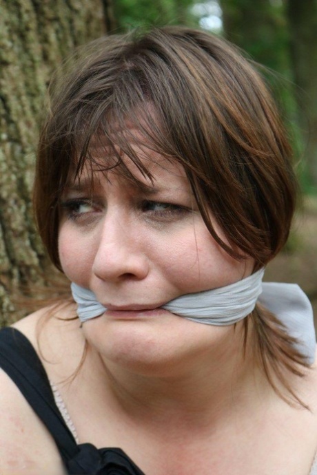 Fat female is gagged and tied to a tree in the woods with her clothes on