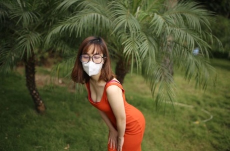 Masked Asian model with glasses posing in her tight orange dress