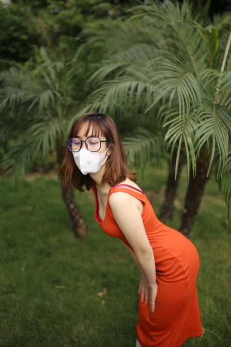 Masked Asian model with glasses posing in her tight orange dress